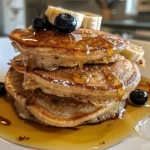 Close-up of golden, fluffy 2 Ingredient Banana Pancakes stacked on a white plate, with banana slices on top.