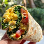 A side view of a breakfast wrap filled with eggs, spinach, and colorful vegetables.