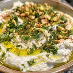 A close-up view of a creamy whipped herb feta topped with chopped pistachios and herbs.
