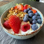 Close-up view of a Greek yogurt breakfast bowl topped with fresh berries, granola, nuts, and a drizzle of honey.
