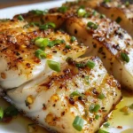 ['Close-up of juicy Garlic Butter Cod Fish fillets garnished with green onions.', 'Side view of succulent cod fish fillets in garlic butter sauce with lemon zest.', 'Deliciously prepared cod fish with a golden garlic butter glaze and green onion topping.', 'Appetizing Garlic Butter Cod Fish fillets displayed on a white plate.']