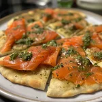 ['A close-up side view of a Smoked Salmon and Pesto Flatbread on a wooden board.', 'Juicy Smoked Salmon and Pesto Flatbread with fresh parsley, captured from the side.', 'Detailed view of a flatbread topped with smoked salmon, pesto, and herbs.', 'Close-up of a delicious-looking Smoked Salmon and Pesto Flatbread garnished with parsley.']