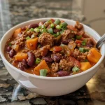 A close-up view of a bowl of beef and sweet potato chili garnished with herbs.